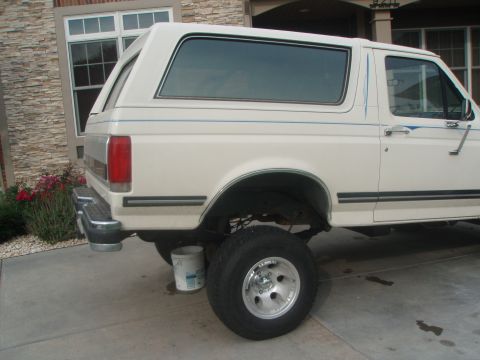 1987 Ford Bronco - The rear passenger side of a white Ford Bronco is shown parked on a concrete driveway next to a house. A white bucket is visible under the…