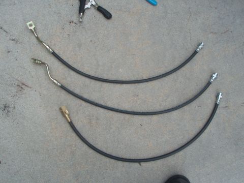 1987 Ford Bronco - Three black rubber brake hoses with metal fittings are laid out on a concrete surface. A pair of pliers is visible in the upper left corner.