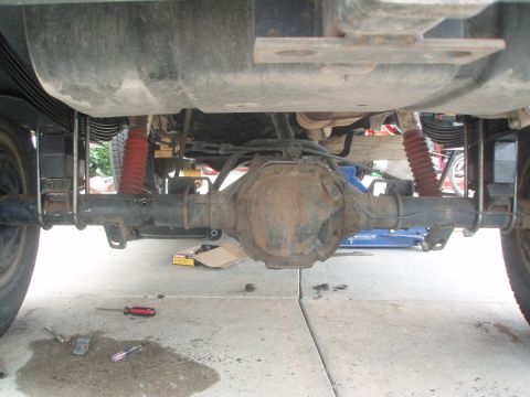 1987 Ford Bronco - The underside of a vehicle, likely a Ford Bronco, showing the rear differential, leaf springs, and red shock absorbers. Tools and a spill a…