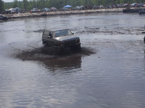 1989 Ford Bronco - A dark-colored SUV is driving through a muddy track, splashing mud as it goes. Several other vehicles and tents are visible in the backgrou…