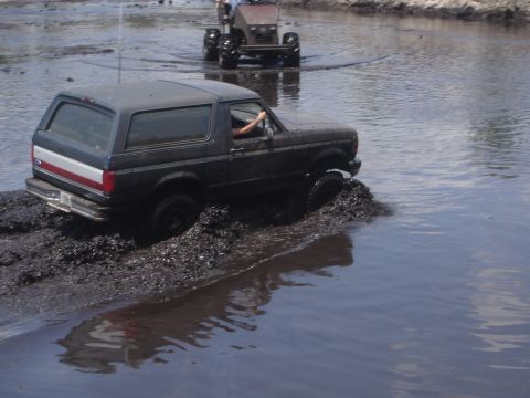 1989 Ford Bronco - A dark-colored SUV is driving through a muddy body of water, with its tires kicking up mud. Another vehicle is visible in the background, a…