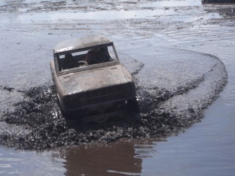 1989 Ford Bronco - A muddy, older model SUV is driving through deep mud and water, kicking up a large spray. The vehicle appears to be a Ford Bronco with its…