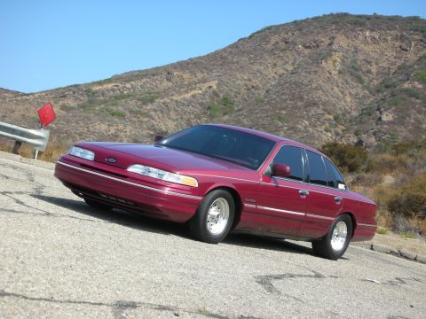 1992 Ford Crown Victoria - A maroon 1992 Ford Crown Victoria is parked on a paved road with a steep, dry, grassy hillside in the background.