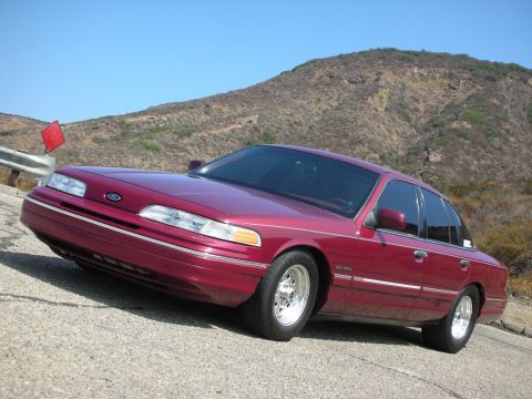 1992 Ford Crown Victoria - A maroon 1992 Ford Crown Victoria is parked on a paved road with a hilly, chaparral-covered landscape in the background under a clear blue…