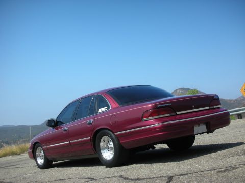 1992 Ford Crown Victoria - The rear passenger side of a dark red sedan is shown parked on a paved road with a mountainous landscape and clear blue sky in the backgrou…