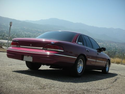 1992 Ford Crown Victoria - The rear passenger side of a dark red 1990s Ford Crown Victoria is shown parked on a gravel road with mountains in the background. The car…