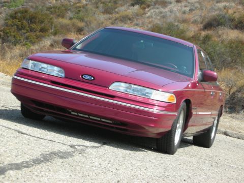 1992 Ford Crown Victoria - A low-angle shot of the front of a maroon 1992 Ford Crown Victoria parked on a gravel road with dry brush in the background.