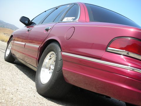 1992 Ford Crown Victoria - A low-angle, side view of a dark red Ford Crown Victoria sedan parked on a gravel shoulder with a clear blue sky overhead. The car features…