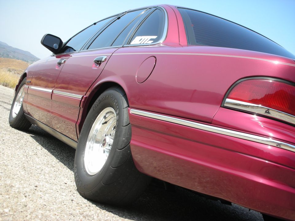 Dark red Ford Crown Victoria sedan with chrome wheels and drag tires, side view.