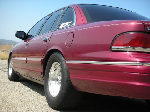 1992 Ford Crown Victoria - The rear passenger side of a modified maroon sedan is shown from a low angle, highlighting its wide drag racing tires and chrome wheels. A…