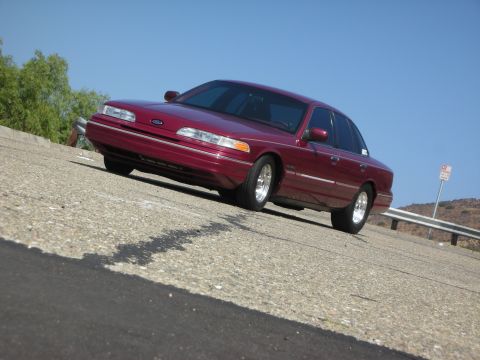 1992 Ford Crown Victoria - A maroon 1992 Ford Crown Victoria is parked on a sloped asphalt road with a clear blue sky above. The car is viewed from a low angle, showi…