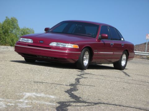 1992 Ford Crown Victoria - A maroon 1992 Ford Crown Victoria is parked on a cracked asphalt road with a clear blue sky in the background. The car is viewed from a low…