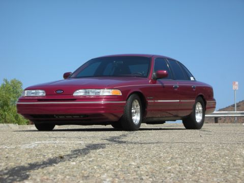 1992 Ford Crown Victoria - A maroon 1992 Ford Crown Victoria is parked on a paved surface with a clear blue sky in the background. The car is viewed from a low front-…
