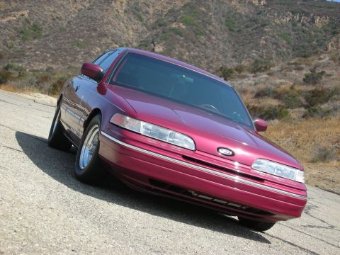 1992 Ford Crown Victoria - A maroon 1992 Ford Crown Victoria is parked on a paved road with a dry, hilly landscape in the background. The car is angled towards the vi…