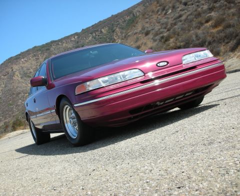 1992 Ford Crown Victoria - A low-angle, front three-quarter view of a dark red sedan parked on a gravel road with a hillside in the background.