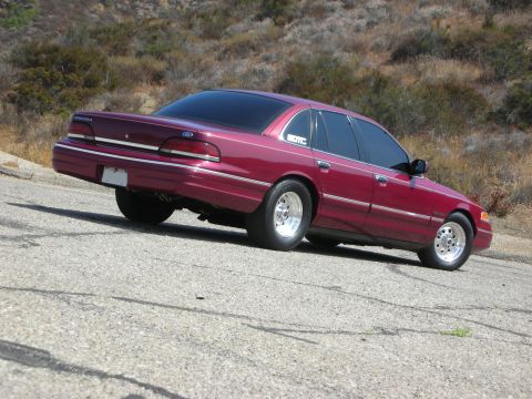 1992 Ford Crown Victoria - A maroon Ford Crown Victoria is parked on a paved road with a dry, hilly landscape in the background. The car has tinted windows and chrome…