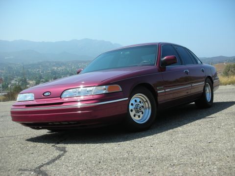 1992 Ford Crown Victoria - Low three-quarter front view of a maroon Ford Crown Victoria sedan on a hilltop asphalt pad, with hazy mountains and a valley of houses beh…