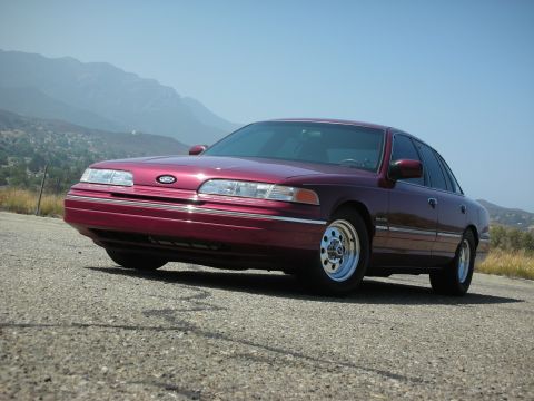 1992 Ford Crown Victoria - A maroon 1992 Ford Crown Victoria sedan is parked on an asphalt road with hills and mountains in the background under a clear blue sky.