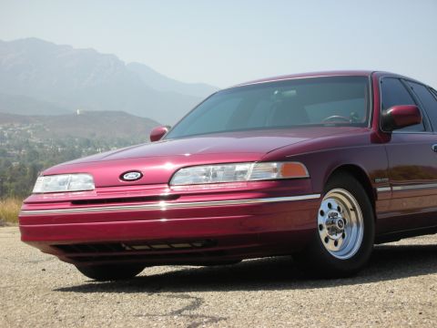 1992 Ford Crown Victoria - A low-angle front three-quarter view of a dark red 1992 Ford Crown Victoria sedan parked on a gravel surface. The car is facing towards the…