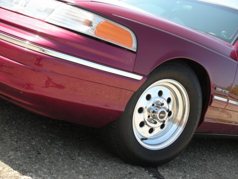 1992 Ford Crown Victoria - Close-up, low-angle shot of the front passenger side of a dark red 1990s Ford Crown Victoria. The image highlights the chrome alloy wheel,…