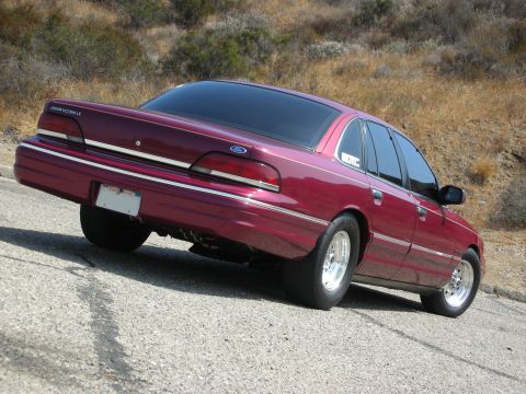 1992 Ford Crown Victoria - The rear driver's side quarter view of a modified maroon 1992 Ford Crown Victoria LX is shown parked on a paved road with a dry, grassy hil…