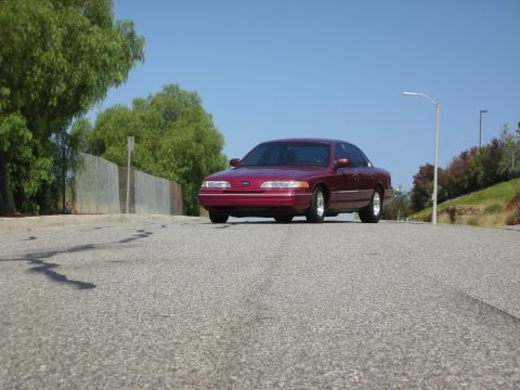 1992 Ford Crown Victoria - A maroon 1992 Ford Crown Victoria is parked on the side of a road with a chain-link fence and trees in the background.