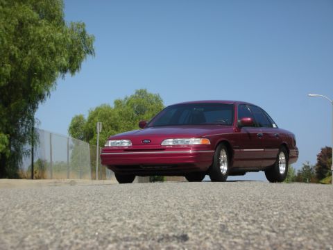 1992 Ford Crown Victoria - A maroon 1992 Ford Crown Victoria is parked on an asphalt road with a chain-link fence and trees in the background under a clear blue sky.