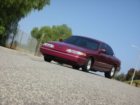 1992 Ford Crown Victoria - A low-angle, wide shot of a maroon Ford Crown Victoria sedan parked on a paved road with a chain-link fence and trees in the background. Th…