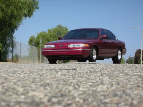 1992 Ford Crown Victoria - A maroon 1992 Ford Crown Victoria is parked on a paved surface with trees and a chain-link fence in the background under a clear blue sky.