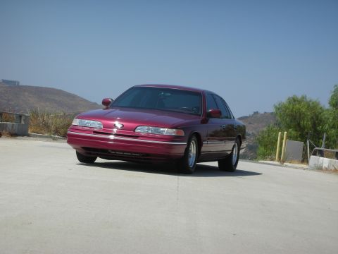 1992 Ford Crown Victoria - A maroon 1990s Ford Crown Victoria sedan is parked on a paved surface with a dry, hilly landscape in the background under a clear blue sky.
