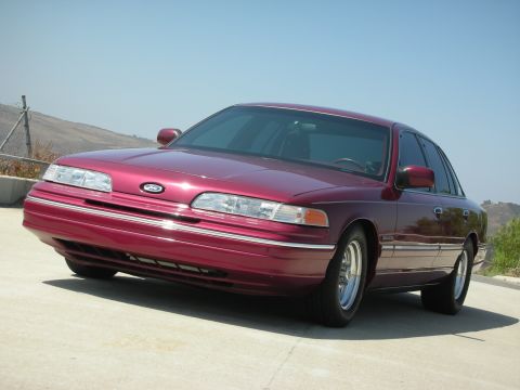 1992 Ford Crown Victoria - A low-angle front 3/4 view of a dark red 1992 Ford Crown Victoria sedan parked on a concrete surface with a clear blue sky and hills in the…
