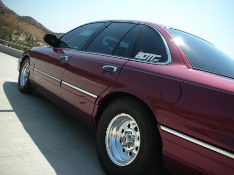 1992 Ford Crown Victoria - The right side of a dark red 1992 Ford Crown Victoria is shown from a low angle, parked on a concrete surface with a mountainous landscape…