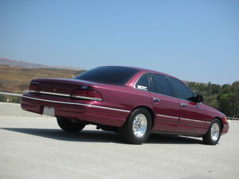 1992 Ford Crown Victoria - A maroon 1992 Ford Crown Victoria is parked on a concrete surface with a clear blue sky in the background. The car is viewed from the rear…