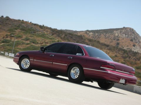 1992 Ford Crown Victoria - A maroon 1992 Ford Crown Victoria LX is parked on a paved surface with a hilly, dry landscape in the background under a clear blue sky. The…