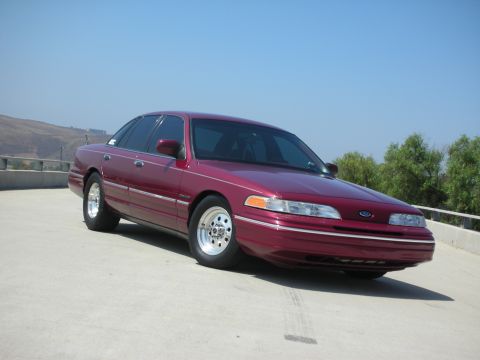 1992 Ford Crown Victoria - A maroon 1992 Ford Crown Victoria is parked on a concrete surface with a clear blue sky overhead. The car is shown from a three-quarter fro…