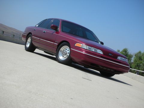 1992 Ford Crown Victoria - A maroon 1992 Ford Crown Victoria is parked on a concrete surface with a clear blue sky in the background. The car is viewed from a low ang…
