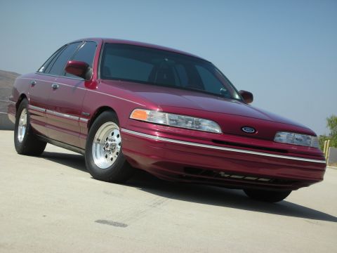 1992 Ford Crown Victoria - A dark red 1992 Ford Crown Victoria is parked on a concrete surface with a clear blue sky in the background. The car is viewed from a low a…