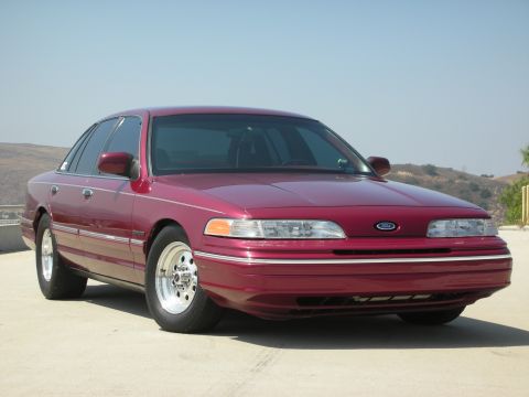 1992 Ford Crown Victoria - A maroon 1992 Ford Crown Victoria is parked on a concrete surface with a hilly landscape and clear blue sky in the background. The sedan fe…