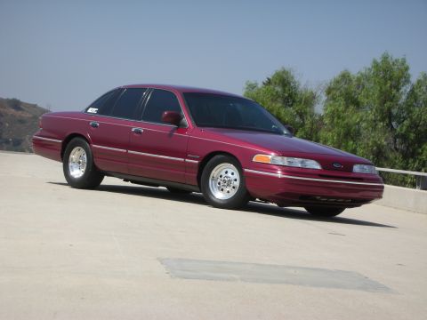 1992 Ford Crown Victoria - A maroon 1992 Ford Crown Victoria is parked on a concrete surface with trees and hills in the background. The car has tinted windows and ch…