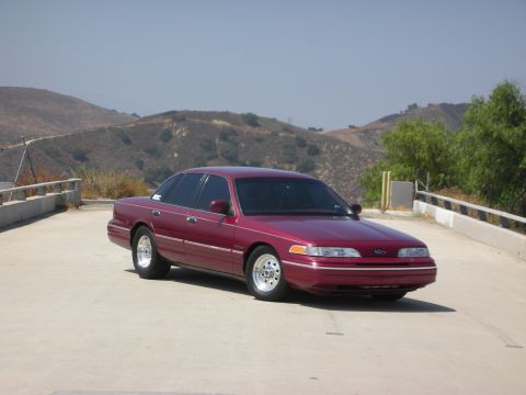 1992 Ford Crown Victoria - A maroon 1992 Ford Crown Victoria sedan is parked on a concrete surface with hills in the background under a clear sky.