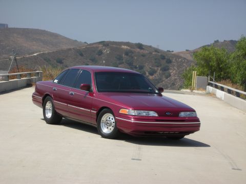 1992 Ford Crown Victoria - A maroon 1992 Ford Crown Victoria is parked on a paved surface with a hilly, dry landscape in the background. The sedan is viewed from a fr…