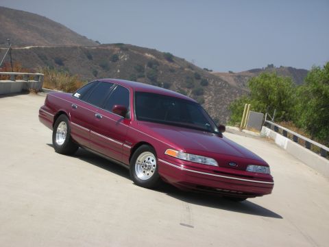 1992 Ford Crown Victoria - A maroon 1992 Ford Crown Victoria is parked on a paved surface with a hilly, arid landscape in the background. The sedan has chrome wheels…
