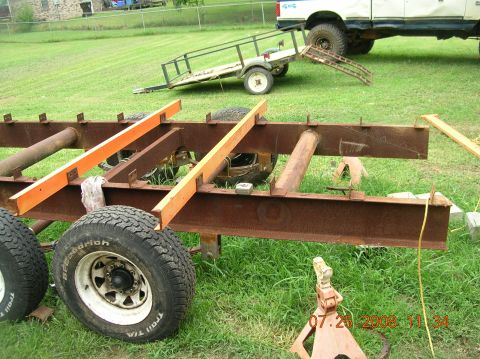1991 Ford F-150 - A trailer frame with dual axles and wheels sits on grass, with a white vehicle in the background.