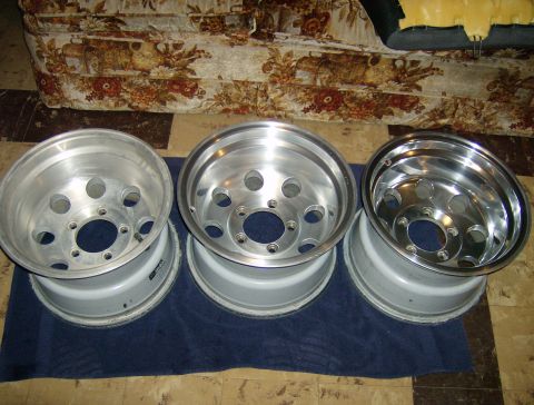 1990 Ford Bronco - Three aluminum alloy wheels are displayed on a dark blue cloth on the floor. The wheels have a five-lug pattern and a polished finish.