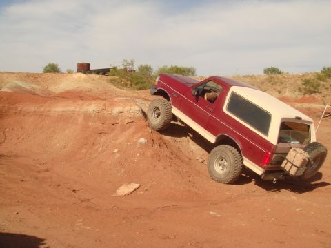 1992 Ford Bronco - A red and tan 1992 Ford Bronco is shown at an angle, climbing a dirt hill. A spare tire and a jerry can are mounted on the rear bumper.