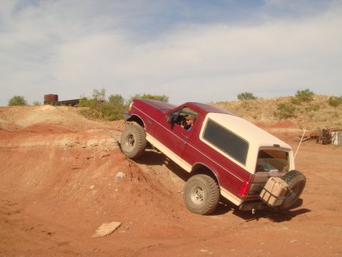 1992 Ford Bronco - A red and cream Ford Bronco is navigating a steep, dusty incline on a dirt trail. The vehicle is angled upwards, with its front wheels elev…