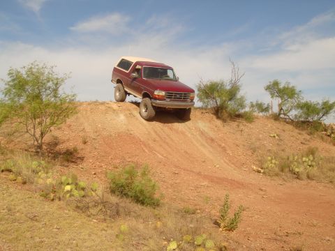1992 Ford Bronco - A red Ford Bronco with a tan camper shell is driving down a steep, dusty hill in a desert environment. The vehicle is angled towards the ri…