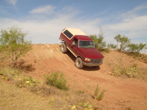 1992 Ford Bronco - A red 1992 Ford Bronco with a tan top is driving up a steep, dusty hill in a desert landscape with scattered vegetation and cacti under a b…
