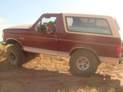 1992 Ford Bronco - A man wearing a baseball cap and sunglasses drives a maroon and tan Ford Bronco off-road. The vehicle is shown from the side, with its larg…