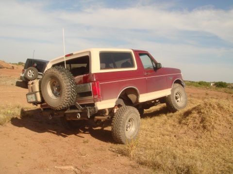 1992 Ford Bronco - A red and tan Ford Bronco is parked on a dirt hill with a black Jeep partially visible behind it. The Bronco has a spare tire mounted on th…